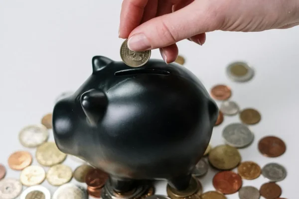 Hand inserting coin into a black piggy bank surrounded by various coins, symbolizing savings.